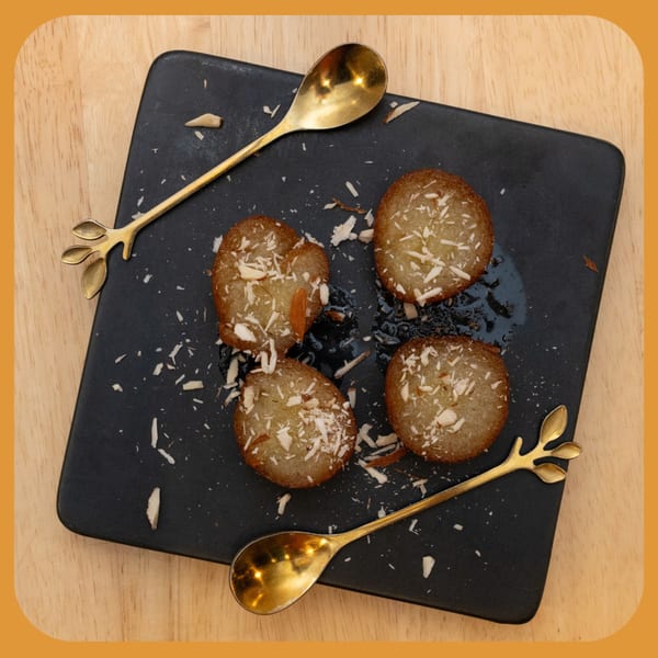 Four Indian Gulab Jamun desserts with shredded coconut and syrup on a black plate with gold spoons.