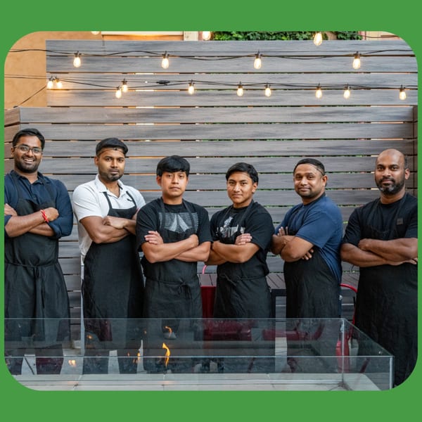 Six male ID Cafe chefs in aprons stand with crossed arms on a patio with string lights.