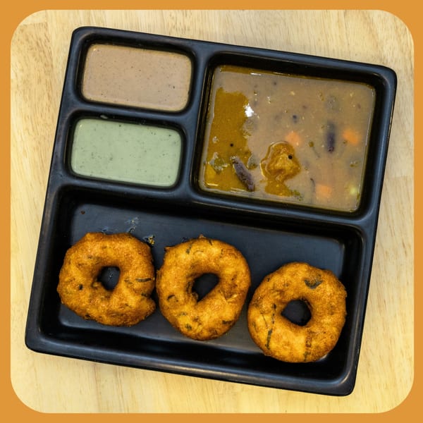 Three golden-brown medu vada, sambar, and two chutneys served on a black tray.